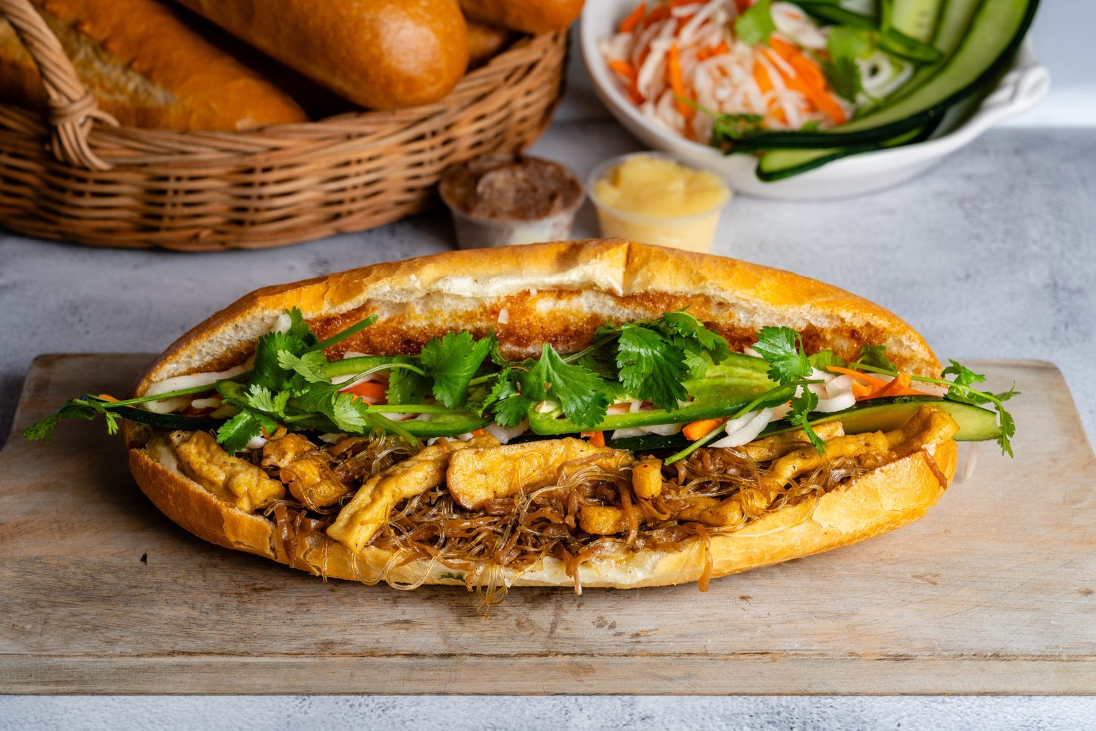 B&aacute;nh m&igrave; sandwich filled with grilled chicken, vermicelli noodles, fresh cilantro, cucumber, and pickled vegetables, served on a wooden board with a basket of bread rolls in the background.