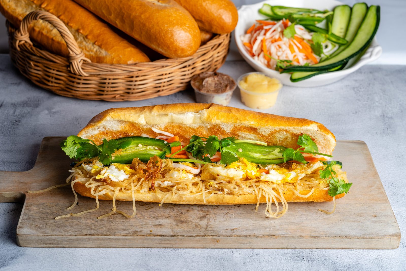 B&aacute;nh m&igrave; sandwich filled with shredded pork skin, fried egg, cucumbers, carrots, and cilantro on a wooden cutting board, with a basket of baguettes and sides of sauces and pickled vegetables in the background.